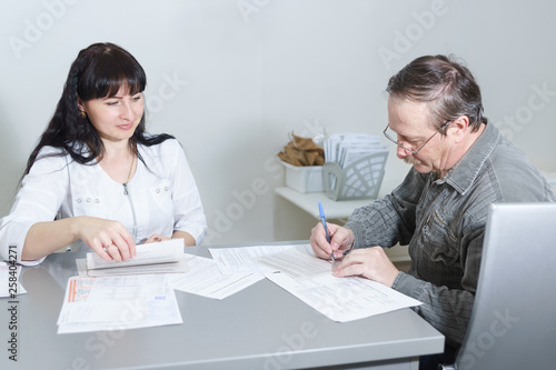 An elderly male patient signs at the reception of a middle-aged woman doctor documents on informed consent to the processing of personal data