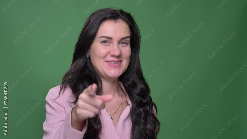 Portrait of young brunette female vloger in pink jacket talking joyfully into camera on green background.