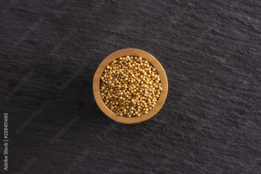 Lot of whole white mustard seeds with wooden bowl copyspace flatlay on grey stone Lot of whole white mustard seeds with wooden bowl copyspace flatlay on grey stone