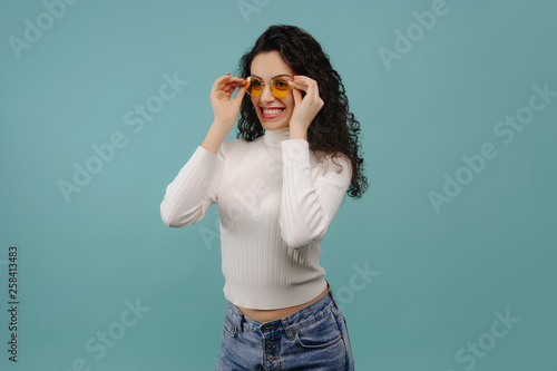 Portrait of smiling beautiful casual girl in yellow sunglasses and blue jeans against of cyan background