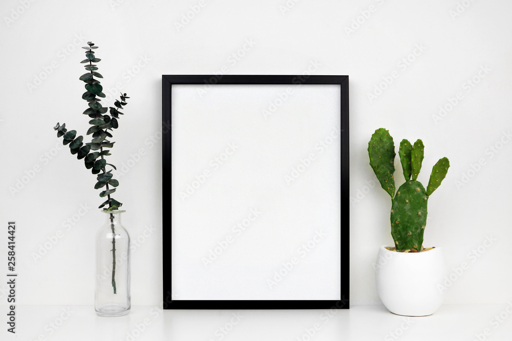 Fototapeta premium Mock up black frame with cactus and branches on a shelf or desk. White shelf and wall. Portrait frame orientation.