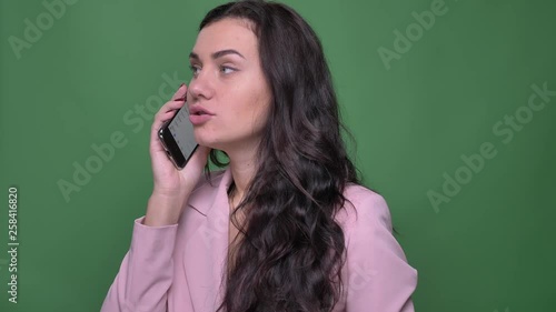 Portrait of young brunette woman in pink jacket talking smilingly on smartphone on green background. photo