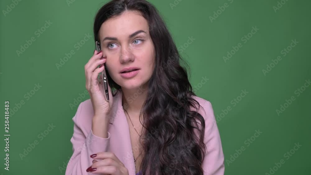 Portrait of young brunette woman in pink jacket talking with friend joyfully on smartphone on green background. Portrait of young brunette woman in pink jacket talking with friend joyfully on smartphone on green background.