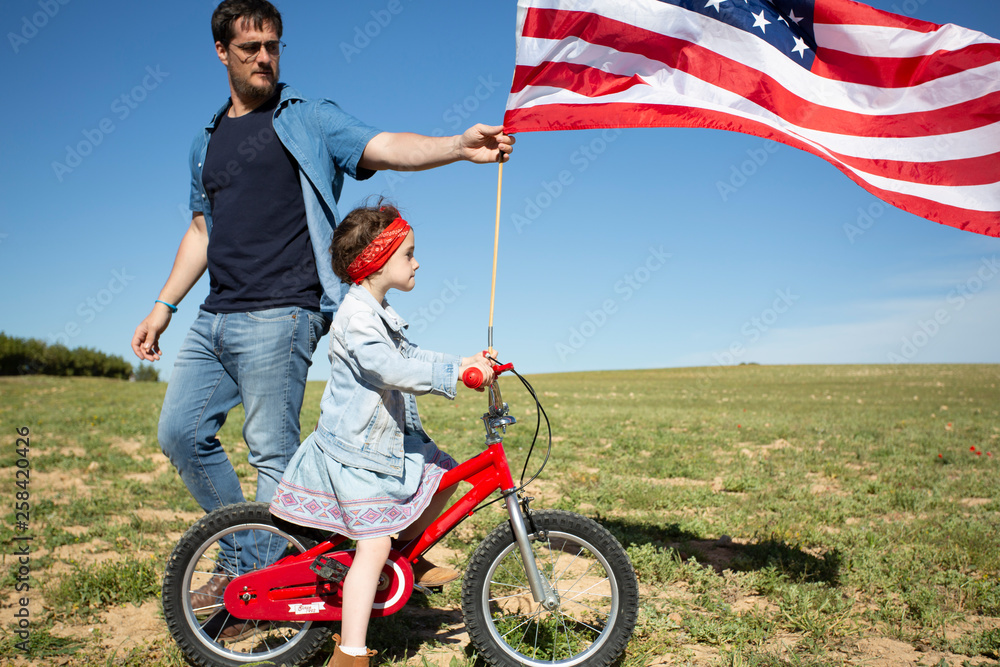 Man and daughter with bicycle and American flag on field in remote landscape