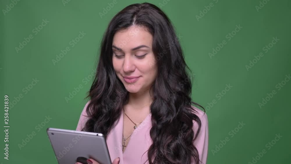 Portrait of young brunette female vloger in pink jacket watching smilingly into tablet on green background. Portrait of young brunette female vloger in pink jacket watching smilingly into tablet on green background.
