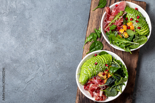 Low carbs bowl. Fresh salad with green spinach, rucola, avocado an ham serrano in white bowl, gray background, top view photo