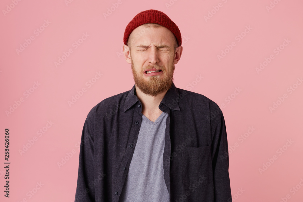 Close up of upset distressed sobbing crying bearded man, with closed eyes, with red hat on the head, in blank shirt and t-shirt, over pink background. Sad emotions concept. Close up of upset distressed sobbing crying bearded man, with closed eyes, with red hat on the head, in blank shirt and t-shirt, over pink background. Sad emotions concept.