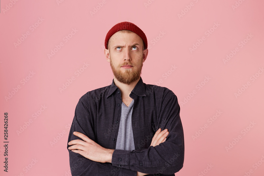 Photo of questioning bearded man in basic shirt, with red hat, with folded arms, look up with one raised eyebrow over pink background. - obrazy, fototapety, plakaty 