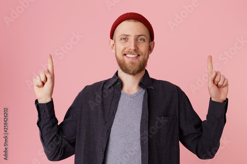 Close up of a happy young bearded man man in basic blue shirt and gray t-shirt, with red hat on the head, smiling and pointing fingers up at copy space isolated over pink background. photo