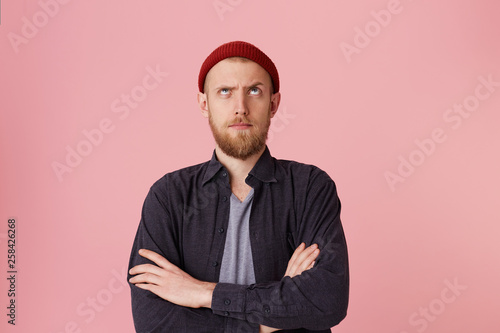 Canvas Print Photo of questioning bearded man in basic shirt, with red hat, with folded arms, look up with one raised eyebrow over pink background