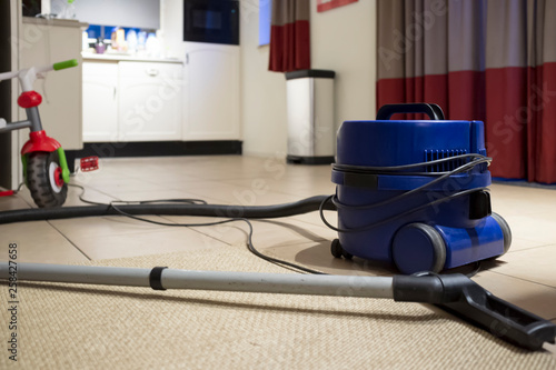 A blue vacuum cleaner stands on the floor after cleaning, next to it is a children's bicycle, against the background of a cozy kitchen. Cleaning in the house.