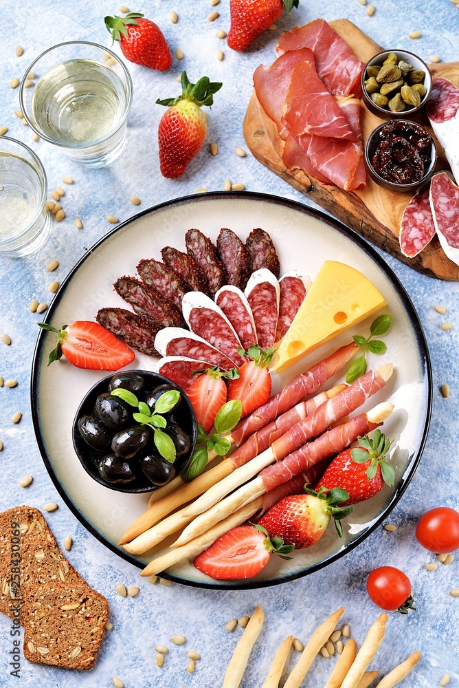Fototapeta premium Antipasti snacks - sausage, homemade grissini with jamon, olives, strawberries, capers, cherry tomatoes, white wine, dried tomatoes and more. Top view.