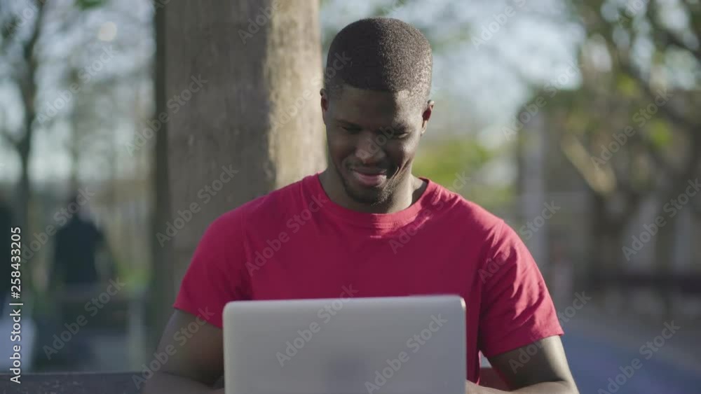 Medium shot of young Afro-American short-cut muscular man in roseT-shirt sitting on bench in park, working on laptop, smiling. Front view. Sun shining. Lifestyle, work concept Medium shot of young Afro-American short-cut muscular man in roseT-shirt sitting on bench in park, working on laptop, smiling. Front view. Sun shining. Lifestyle, work concept