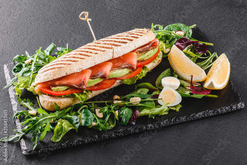 Sandwich with red fish, eggs, avocado, fresh vegetables and greens on black shale board over black stone background. Healthy food concept