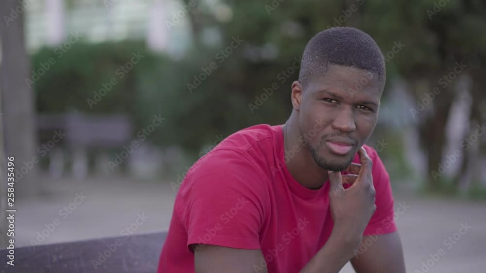 Medium shot of young Afro-American short-cut muscular man in rose T-shirt sitting on bench in park, touching chin, turning head to camera, looking at camera. Dolly shot. Lifestyle concept Medium shot of young Afro-American short-cut muscular man in rose T-shirt sitting on bench in park, touching chin, turning head to camera, looking at camera. Dolly shot. Lifestyle concept