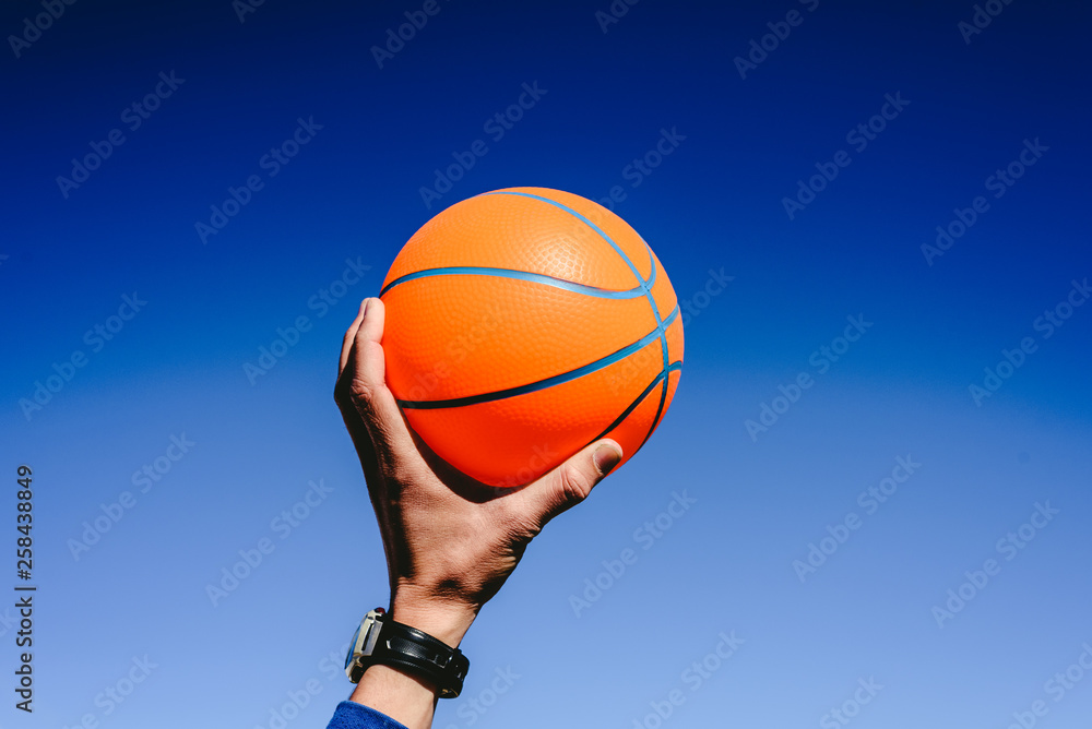 Hand holding an orange basketball ball on blue sky background, invitation to play, copy space free area. Hand holding an orange basketball ball on blue sky background, invitation to play, copy space free area.