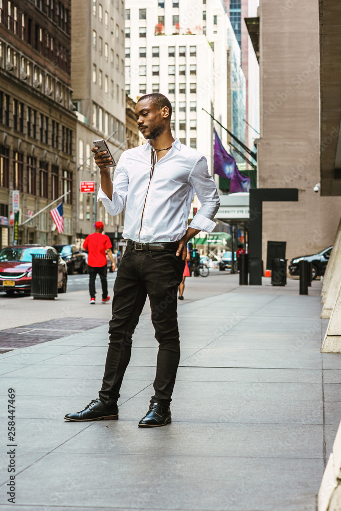 Young African American Man with beard, wearing white shirt, black pants,  leather shoes, standing on street in Manhattan, New York, reading messages  on cell phone. People, cars, buildings on background Stock Photo