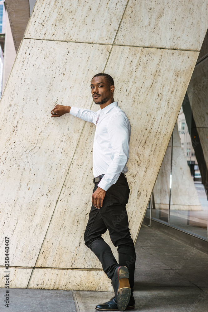 Portrait of Young African American Man in New York City. Black guy with  beard, wearing white long sleeve shirt, black pants, leather shoes,  standing against column outside office building, thinking.. Photos