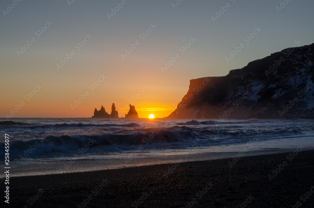 Sunset behind jagged cliffs, over the ocean waves, and black sand beach of Vik Iceland