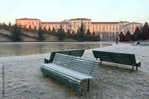 Benches stand in the Park of the Portello district on the grass in the frost on the background of a nursing home - the Palazzolo-Don Gnocchi Institute in Milan, Italy on a winter cloudy morning.