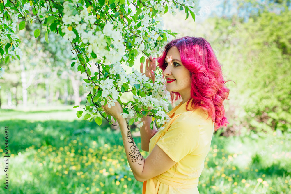 Beautiful and young woman with bright red hair and ref lips standing next to a blooming apple tree in a yellow dress. Beautiful and young woman with bright red hair and ref lips standing next to a blooming apple tree in a yellow dress.