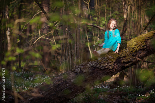 Beautiful little girl in a blue dress walking in the spring wood. Pretty girl sitting on the tumbled-down tree. Kid playing outdoors in summer. Easter time.