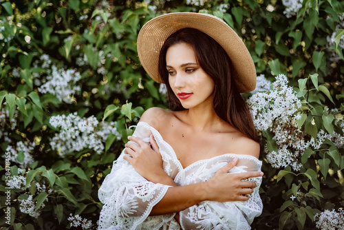 young woman in a straw hat in lilac. Model in white pennuar among flowers