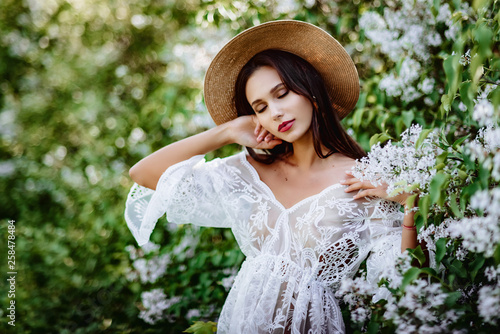 young woman in a straw hat in lilac. Model in white pennuar among flowers