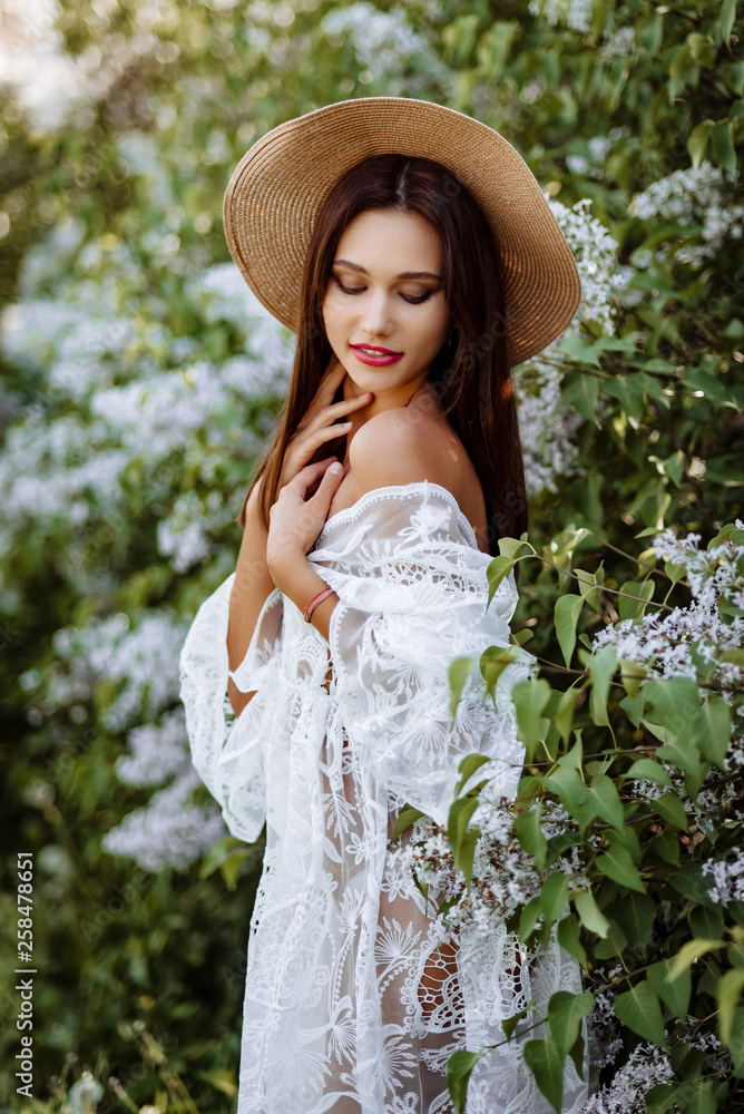 young woman in a straw hat in lilac. Model in white pennuar among flowers