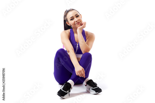 A dark-haired woman coach in a sporty purple short top and gym leggings posing, sitting on floor and smilling on a white isolated background in studio