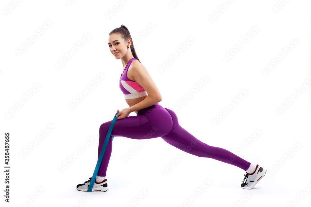 A dark-haired woman coach in a sporty purple short top and gym leggings makes lunges with sport fitness rubber bands on a white isolated background in studio A dark-haired woman coach in a sporty purple short top and gym leggings makes lunges with sport fitness rubber bands on a white isolated background in studio