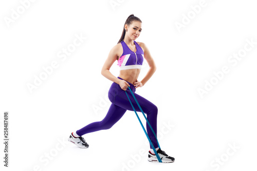 A dark-haired woman coach in a sporty purple  short top and gym leggings makes lunges  with sport fitness rubber bands  on a  white isolated background in studio © Виталий Сова