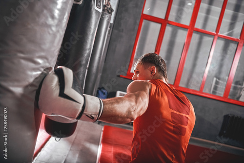 Strong punch. Muscular athlete in white gloves training on heavy punch bag in boxing gym photo