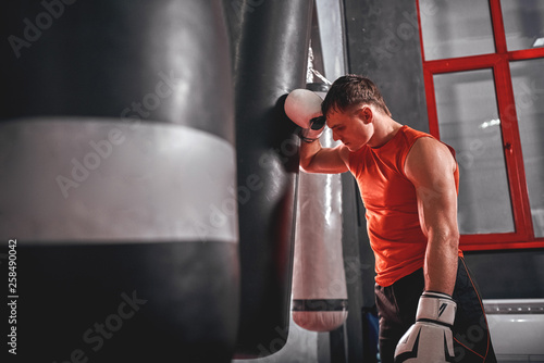 Tired after workout. Tired young athlete in sports clothing looking away after hard training on heavy punch bag in boxing gym photo