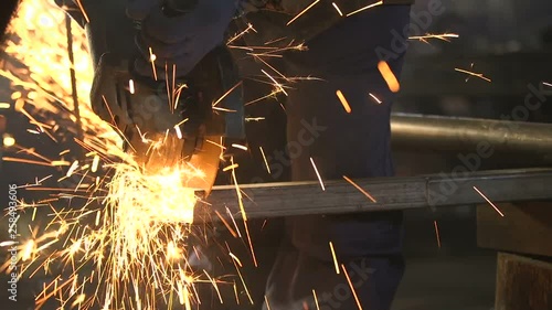 Worker with a side grinder cutting a long metallic tube with yellow sparkles