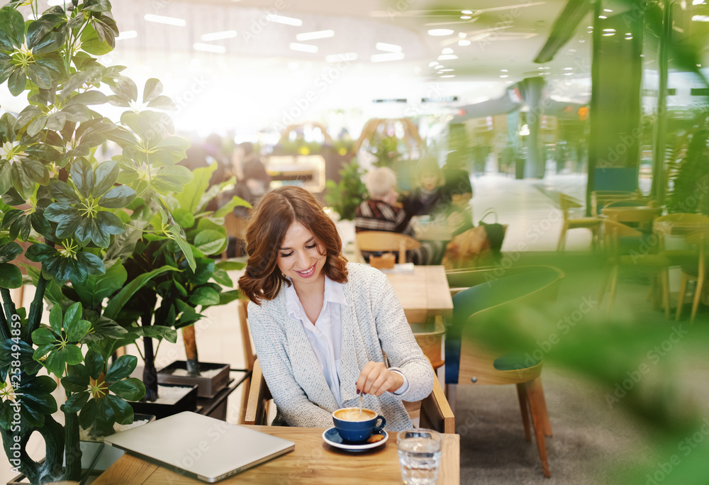 Charming brunette with toothy smile dressed casual stirring coffee while sitting in cafeteria. On desk glass of water and laptop. Charming brunette with toothy smile dressed casual stirring coffee while sitting in cafeteria. On desk glass of water and laptop.