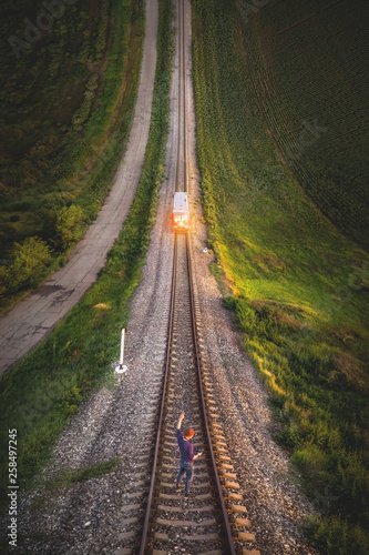 Aerial view A male hipster in a cap holding up a hand with a gesture Victory is standing on rails in front of an approaching train. Reverse vertical panorama in the style of Inception