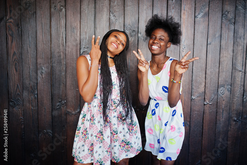 Two black african girlfriends at summer dresses posed against dark wooden background and showing two fingers.