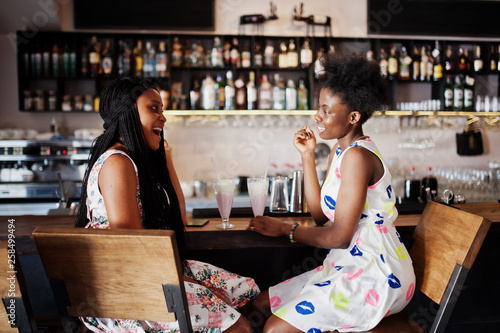 Two black african girlfriends at summer dresses drinking milkshake cocktails in bar.