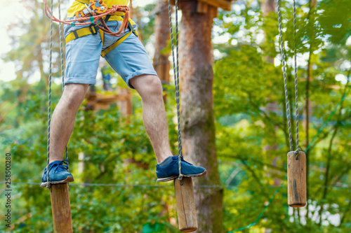Male legs in blue sneakers keep balance on wooden stumps hanging on ropes on high trees in park. Rope park with different obstacles and ziplines. Extreme rest and summer activities concept.