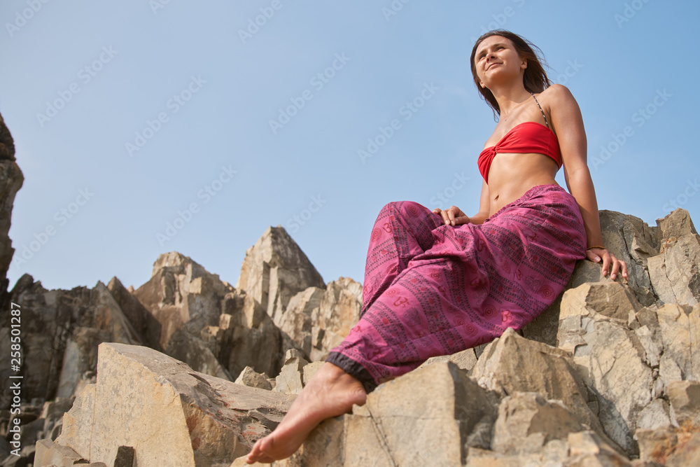 young woman with dark hair in a swimsuit and wide pants sits on a rock against the sky. the concept of rest in the warm season