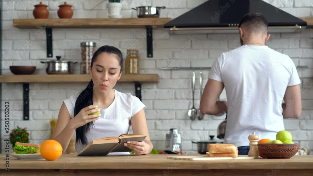 Couple at the kitchen. Breakfast, morning time. Young woman reading book and eating apple. Guy giving cup of coffee to her. Couple at the kitchen. Breakfast, morning time. Young woman reading book and eating apple. Guy giving cup of coffee to her.