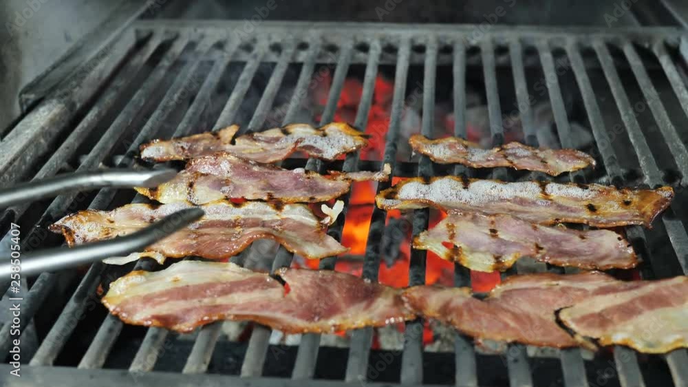 Slices of bacon are grilled, the fat from the bacon is drained from the fried bacon, the chef in black gloves with iron tongs turns over the slices of bacon alternately for the final preparation Slices of bacon are grilled, the fat from the bacon is drained from the fried bacon, the chef in black gloves with iron tongs turns over the slices of bacon alternately for the final preparation