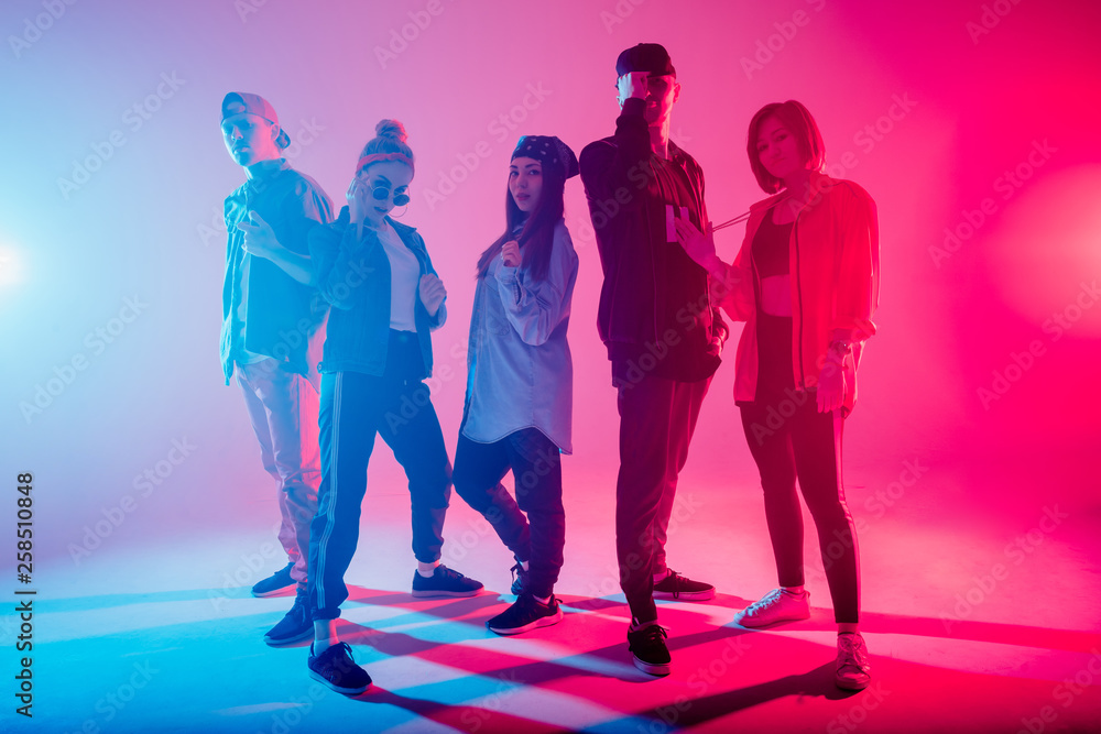 Young modern dancing group of five adult young people practice dancing on colorful background. Fashionably dressed youngsters posing over blurred disco club color lights Young modern dancing group of five adult young people practice dancing on colorful background. Fashionably dressed youngsters posing over blurred disco club color lights