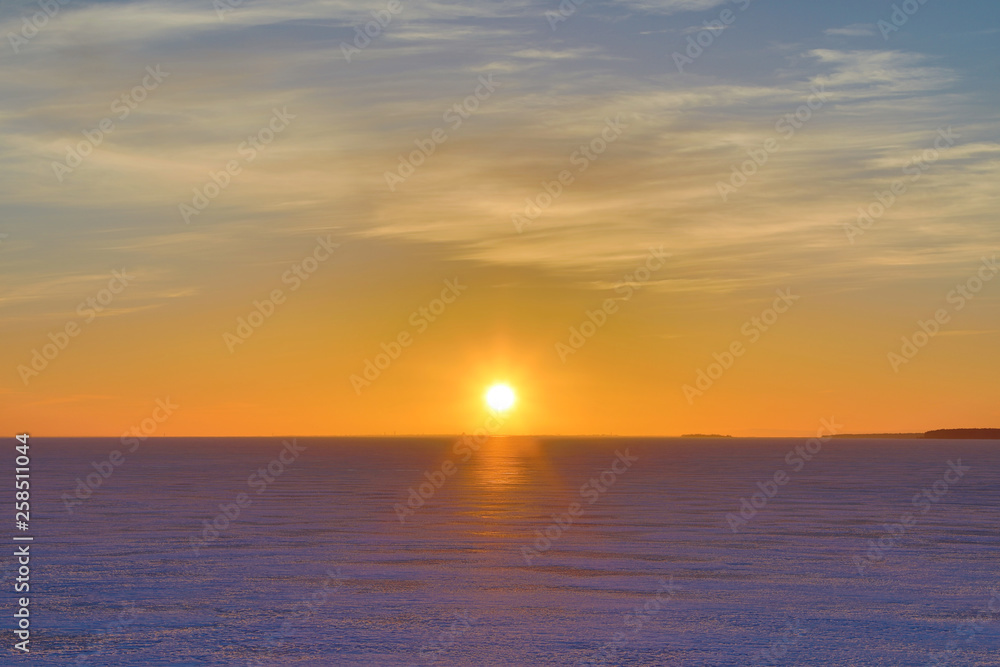 The sunset or sunrise. The cloudy sky cloured in red, orange, crimson, purple, violet and blue bright and vivid coloures with setting or rising sun over the sea, lake or bay covered with ice and snow
