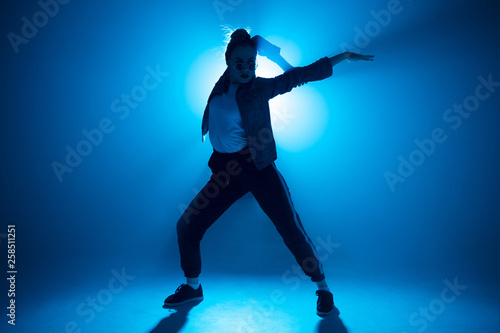 Young woman dressed in street fashion wear dancing hip-hop style over studio blue light background with flare effects © alfa27