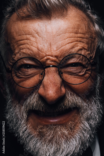 Mature man with angry grimace on face revealing in screwed-up eyes, evil wicked smile and old deep wrinkles close-up. Human emotions and facial expressions concept.