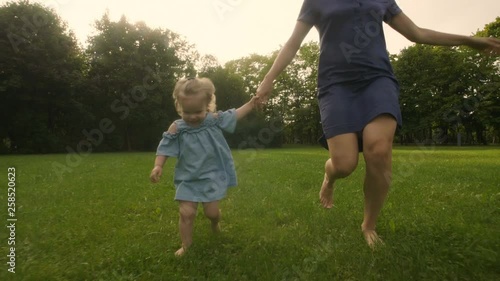 Baby girl with mother running through the grass in the summer sunlight in park. Baby have fun. Slow motion. Happy family concept. 