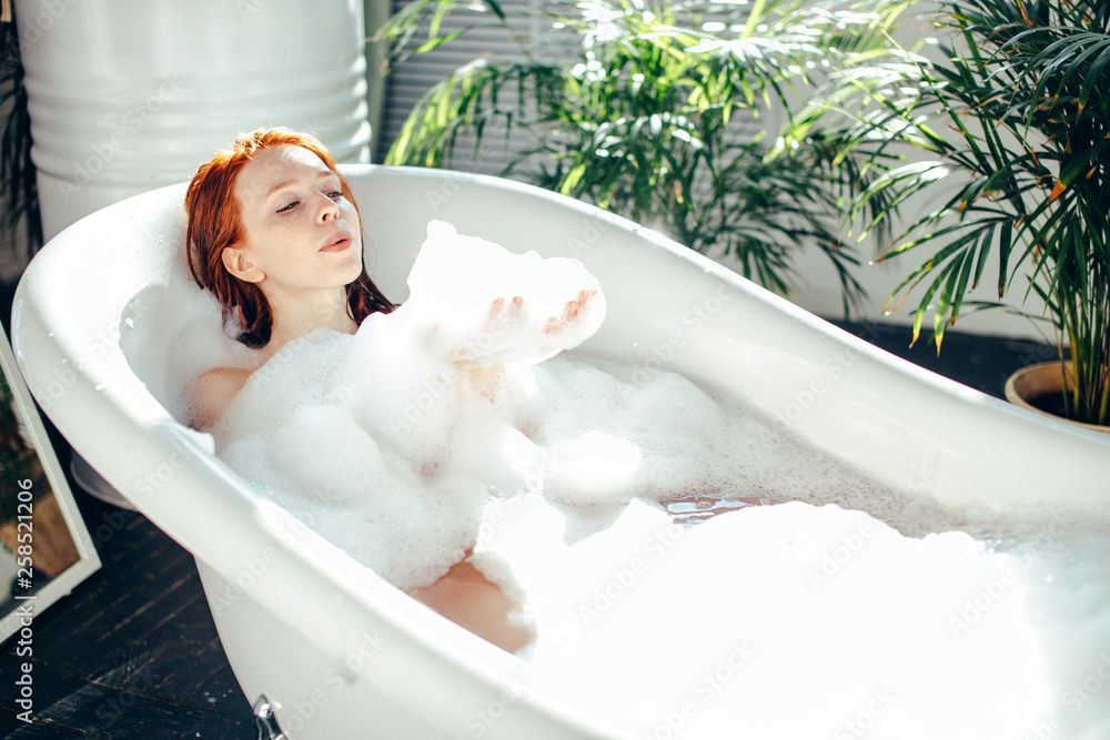 Playful young caucasian woman enjoying foam bath at home. Resting, relaxing, blowing on a cloud of foam - obrazy, fototapety, plakaty 