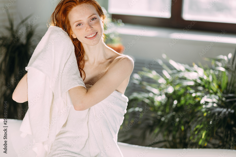 Young redhead beautiful woman beaming with heapiness and joy is sitting on the edge of a bathtub in bathroom and wiping her long ginger hair with towel.